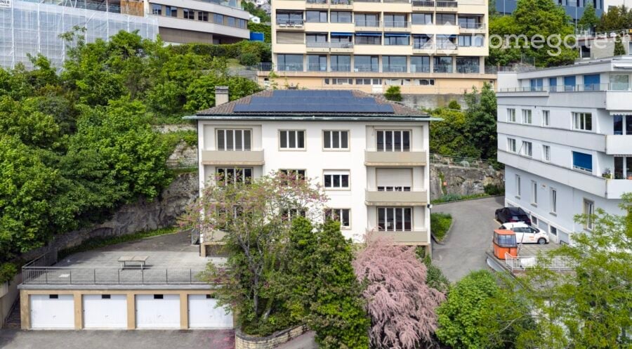 A multi-story residential building with solar panels on the roof, surrounded by greenery and neighboring structures. Features include a garage area with multiple parking spaces.
