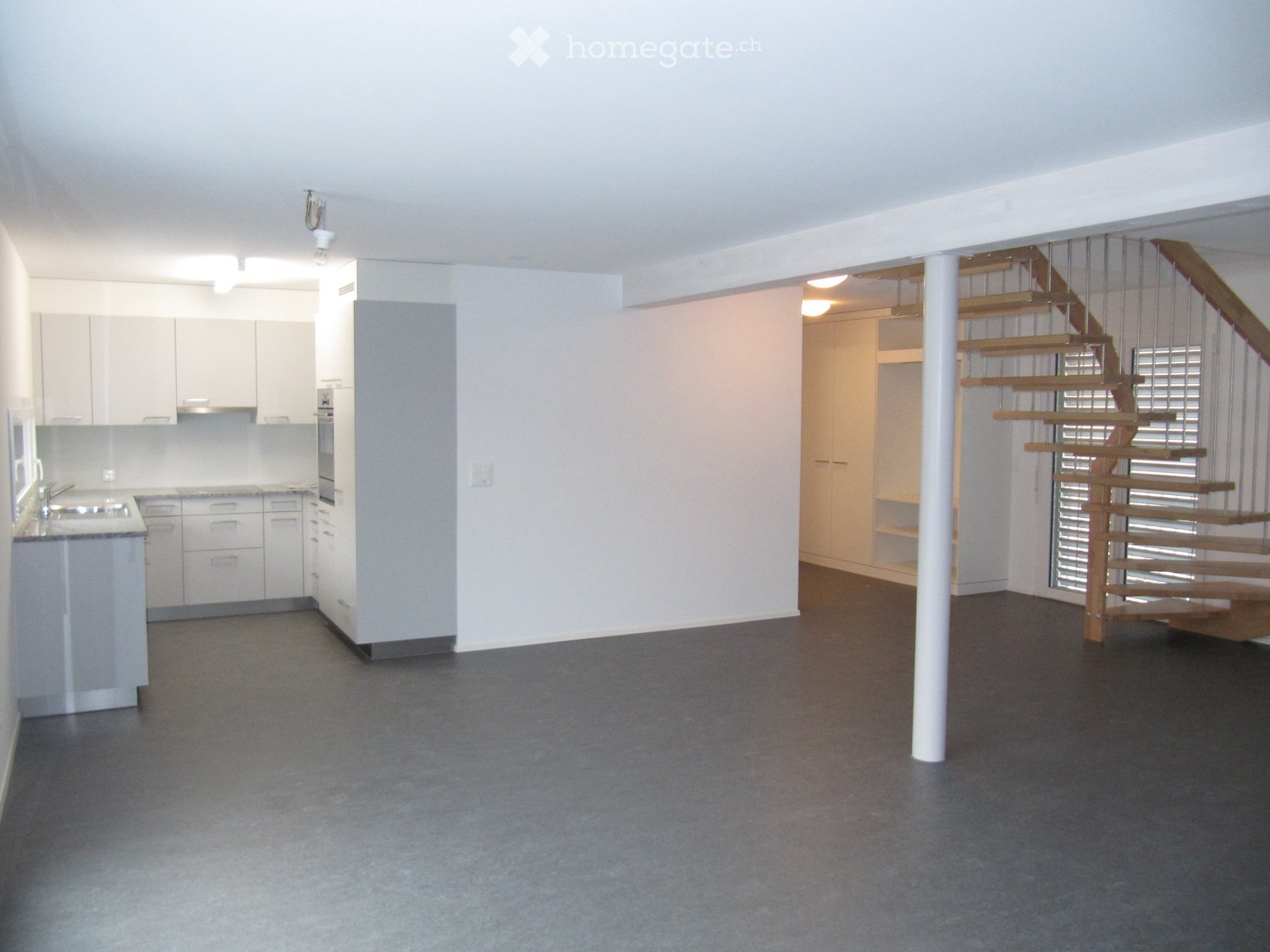 Unfurnished Room. Open-plan space with a modern kitchen area, a staircase leading to another level, and a central support column. The kitchen features white cabinets, a sink, and a countertop. The floor is covered with dark grey carpeting.