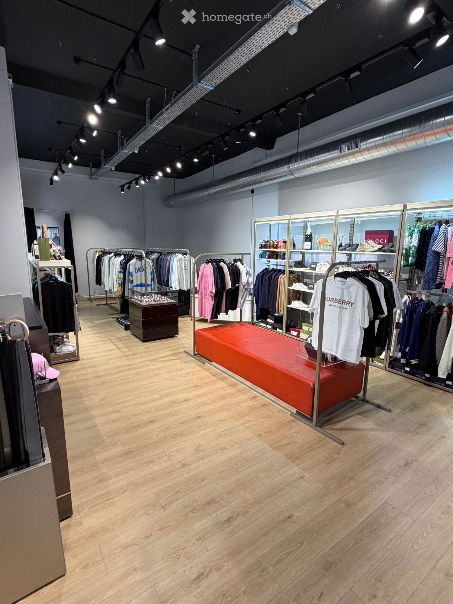 Modern retail space with wooden flooring, clothing racks, a red bench, shelves, and display cases.