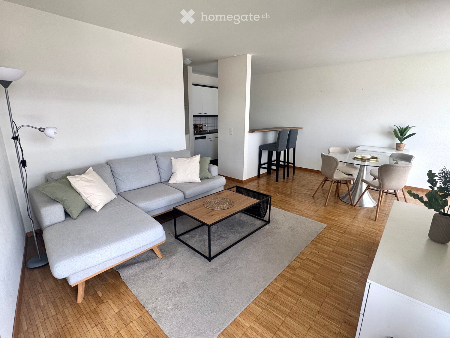 Modern living room with a grey sectional sofa, wooden coffee table, dining area with chairs and table, and a small kitchen area with stools.