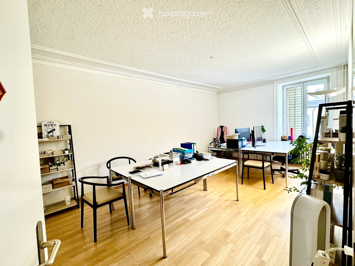 Bright, modern room with wooden flooring, white walls, and large windows. Contains a white table with chairs, a shelf with books and decorative items, a laptop, and a potted plant.