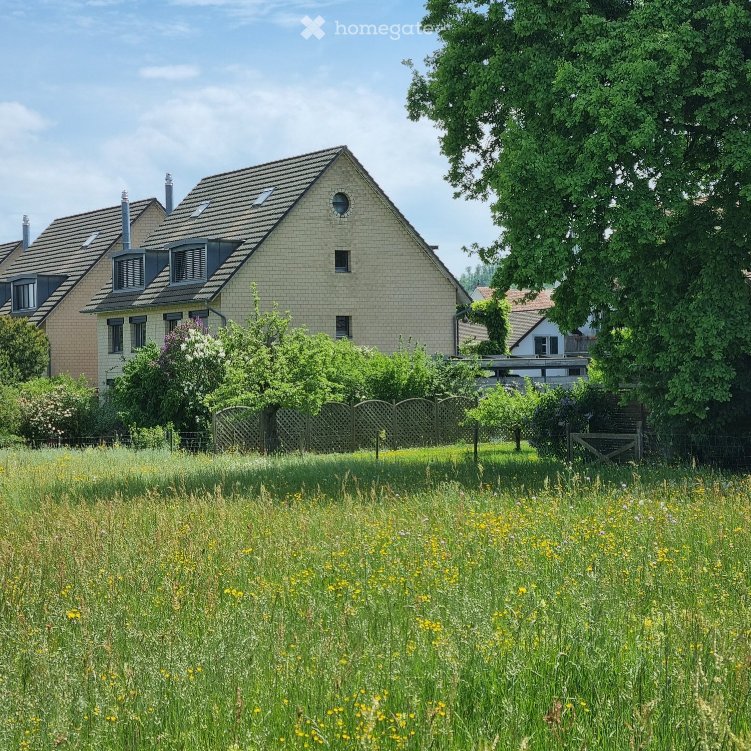 Two-story house with a tiled roof, light green siding, multiple windows, and a fenced garden area with tall grass and wildflowers.