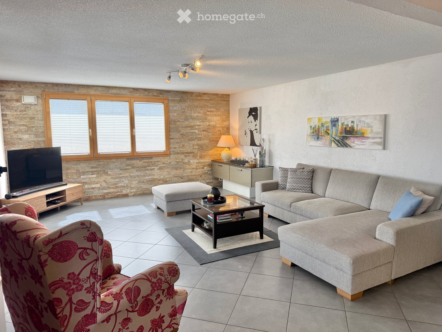 Modern living room with a grey sectional sofa, coffee table, ottoman, armchair, TV stand, table lamp, and artwork on the walls.
