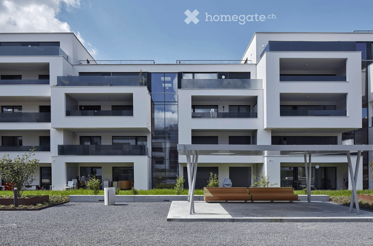 Modern multi-story residential building with white facade, large windows, and balconies. The ground floor features a covered entrance area with seating and greenery.