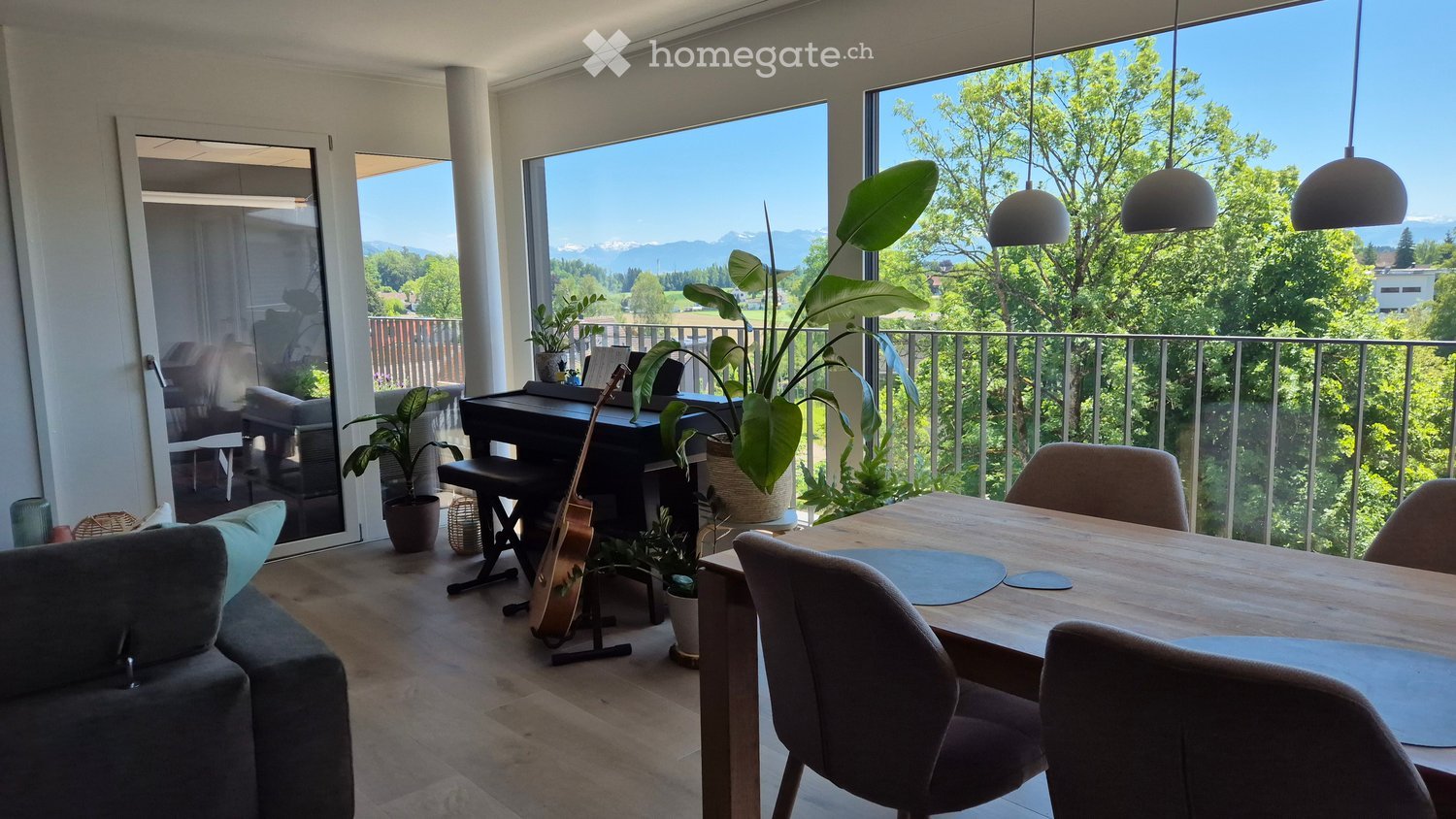 Bright living space with large windows offering a scenic view, furnished with a sofa, dining table, chairs, a guitar, a piano, and potted plants.