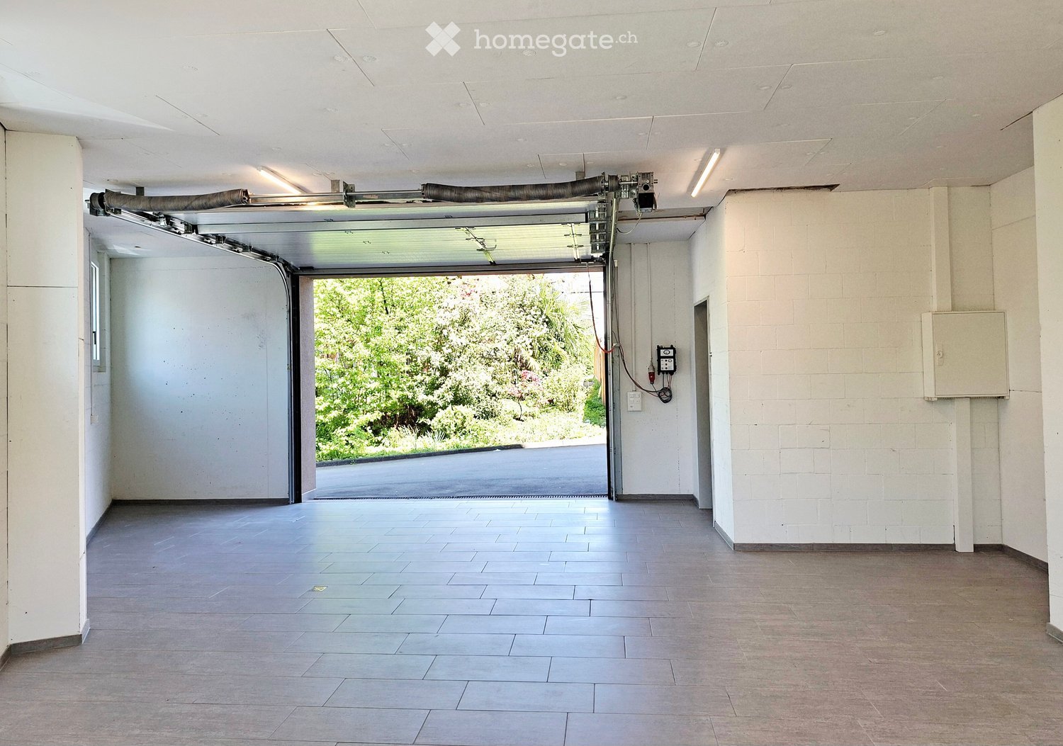 Empty garage with tiled floor, white walls, and an open automatic door leading to a driveway and greenery outside.