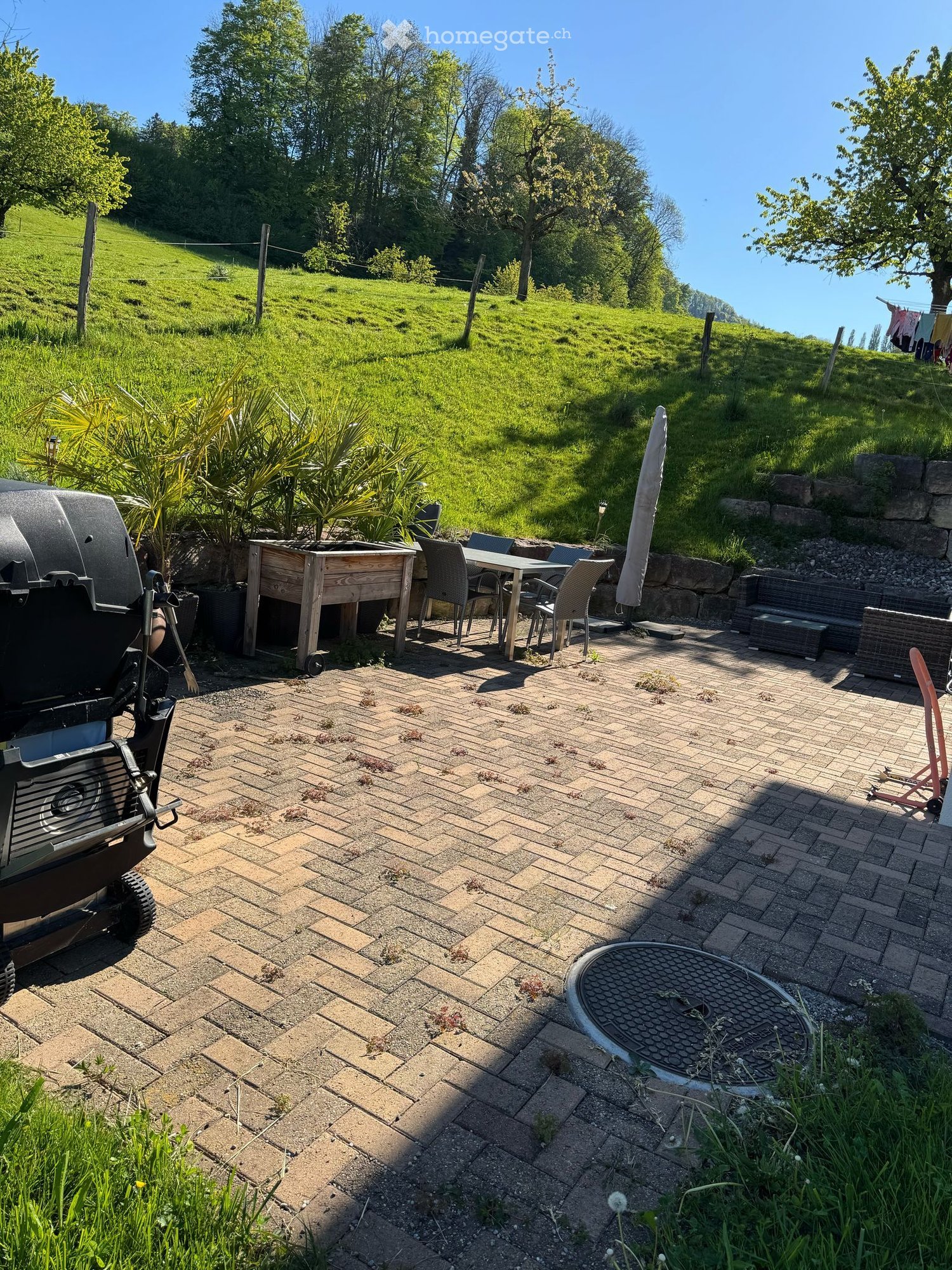 Outdoor patio with brick tiles, a wooden table and chairs, a grill, and a bench, surrounded by greenery and a grassy hill.