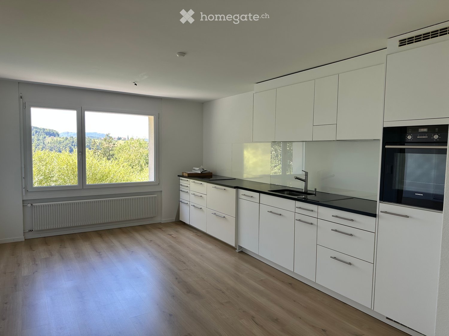 Modern kitchen with white cabinets, black countertops, a sink, a faucet, and an oven. Large windows offer a scenic view of greenery.