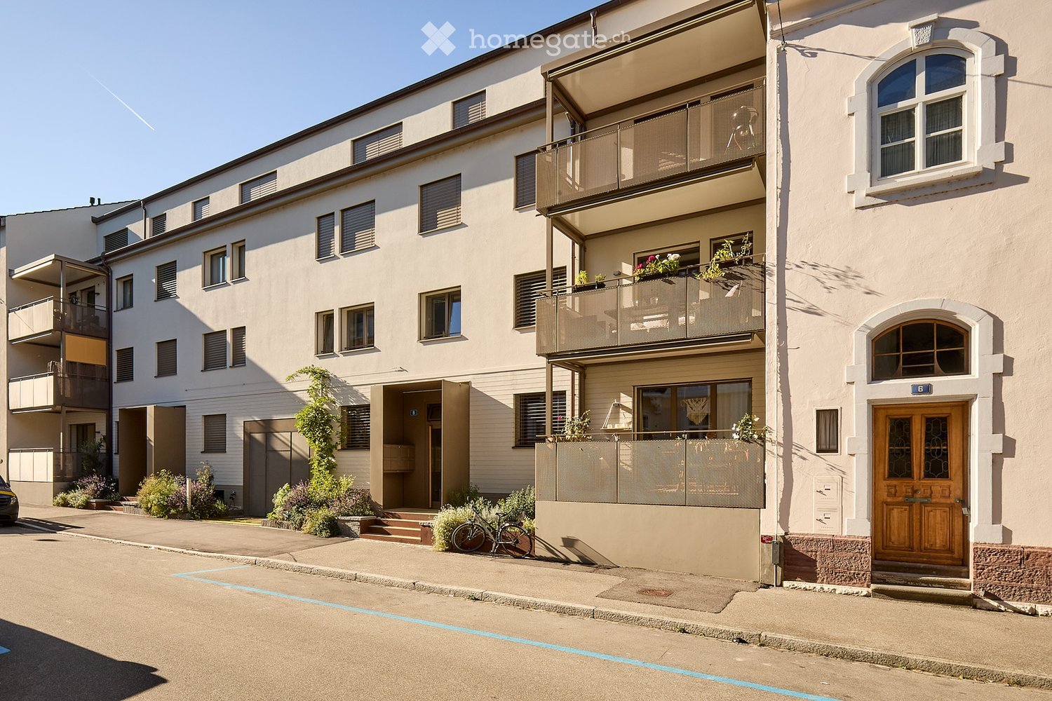 Multi-story residential building with balconies, large windows, and a wooden front door. The building has a light-colored facade and is surrounded by greenery and a paved street.