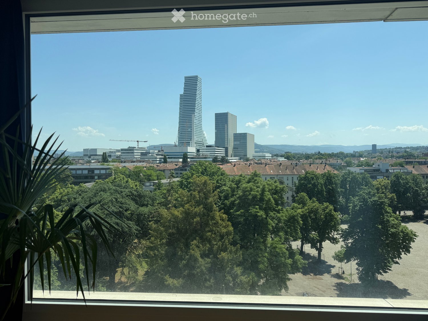 View from a balcony showing a cityscape with modern skyscrapers, trees, and a clear blue sky.