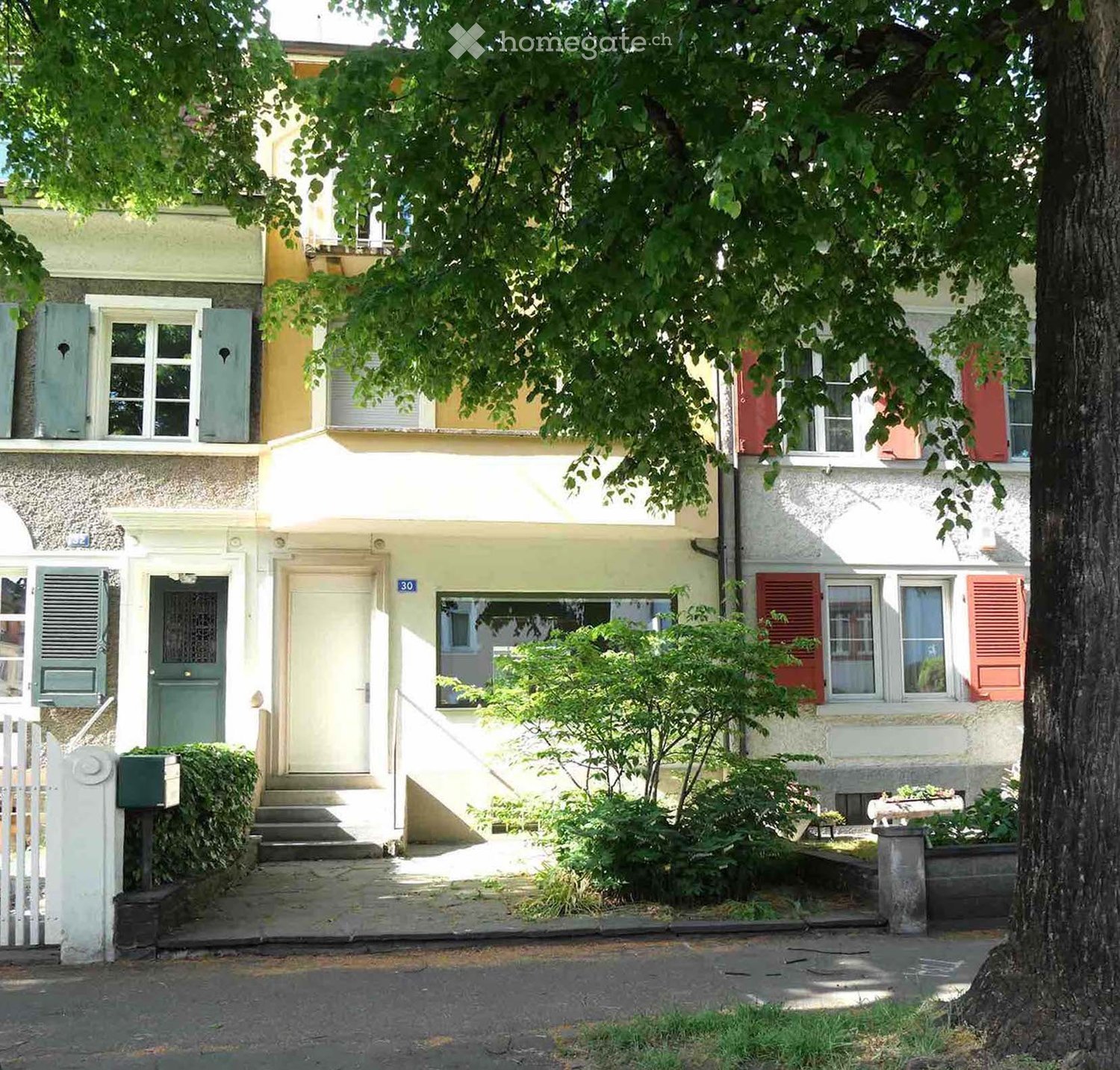 Two-story residential building with light yellow facade, green shutters, and a small garden area with trees and shrubs.