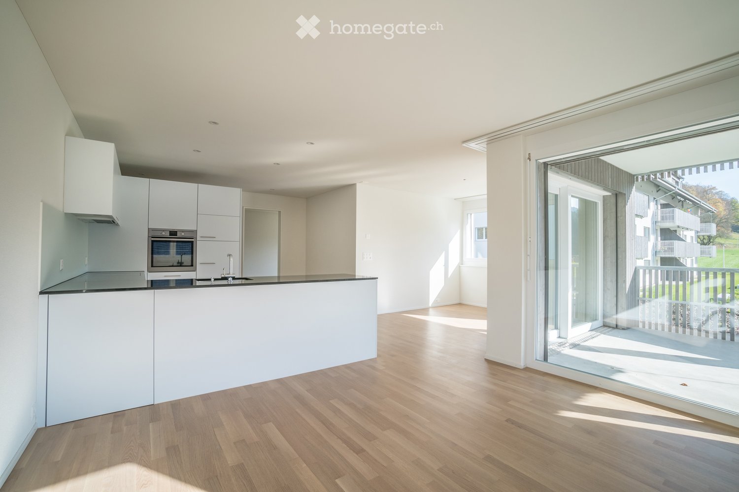 Modern kitchen with white cabinets, a central island, and a large window leading to a balcony. The floor is light wood, and the space is well-lit with recessed lighting.