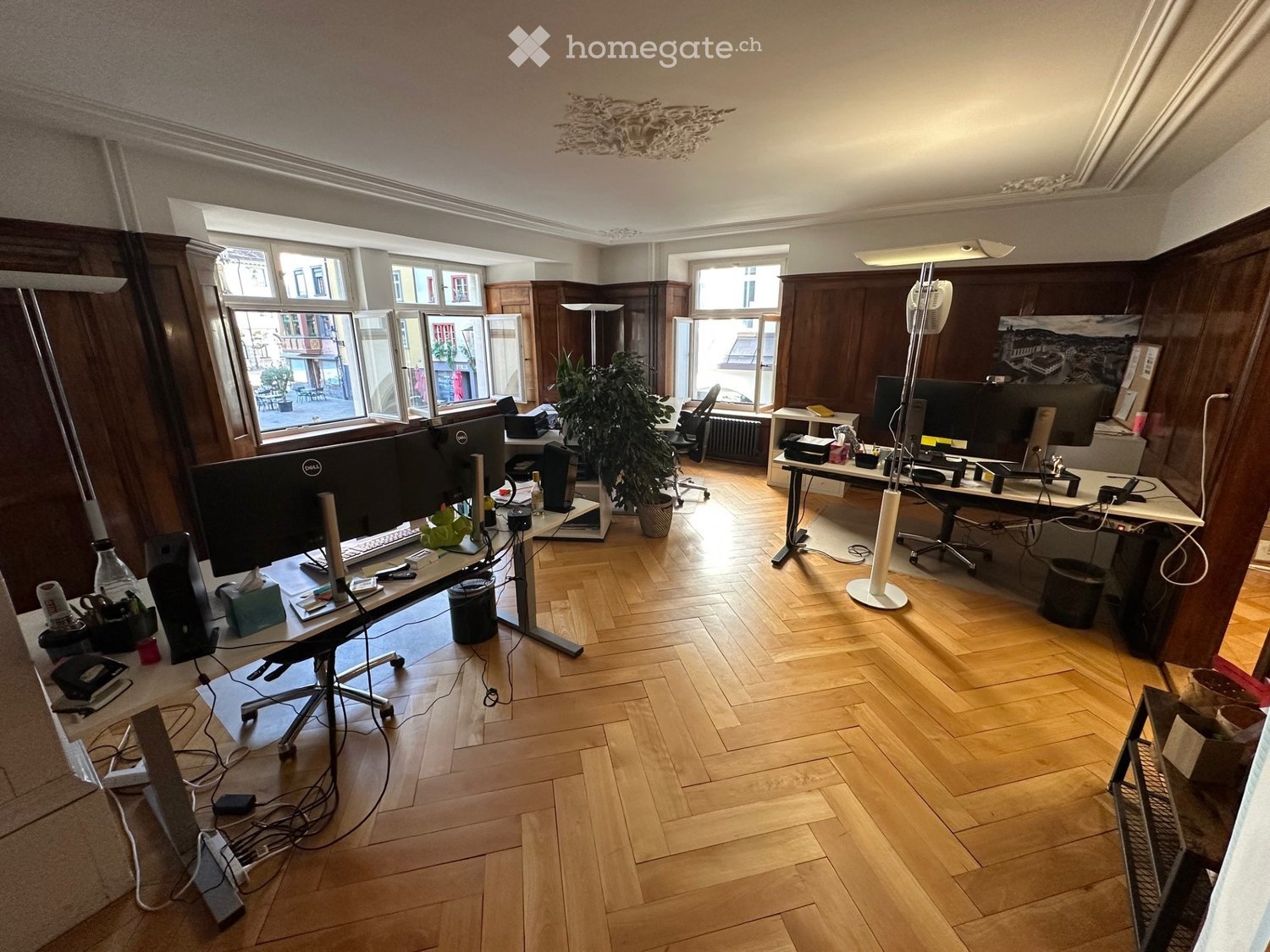 Office space with wooden flooring, large windows, two desks with multiple monitors, chairs, and office supplies.