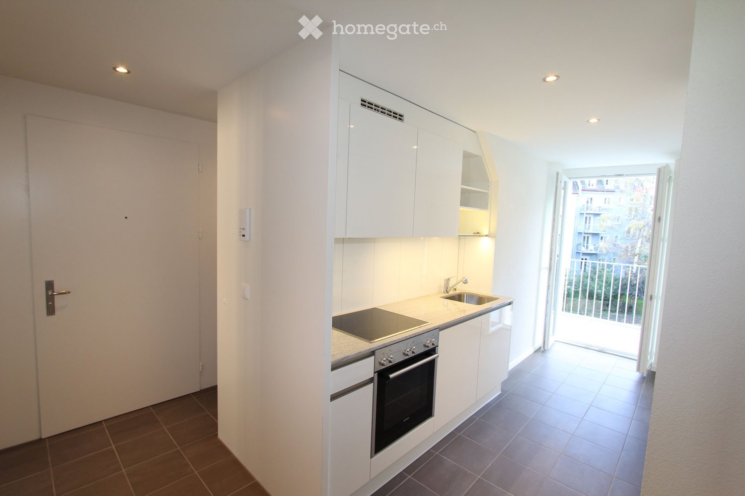 Modern, compact kitchen with white cabinets, an oven, a sink, and a faucet. The space features a tiled floor and an open doorway leading to a balcony.