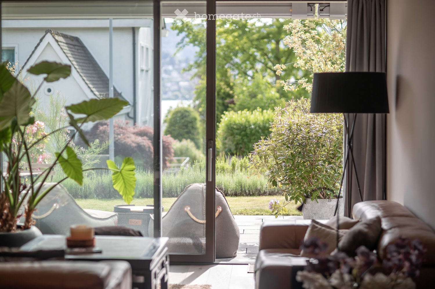 Modern living room with large glass doors opening to a garden, featuring a brown leather sofa, coffee table, floor lamp, and decorative plants.