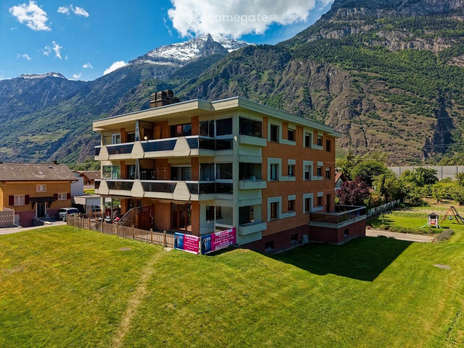 A multi-story residential building with balconies, surrounded by a well-maintained lawn and set against a backdrop of snow-capped mountains.