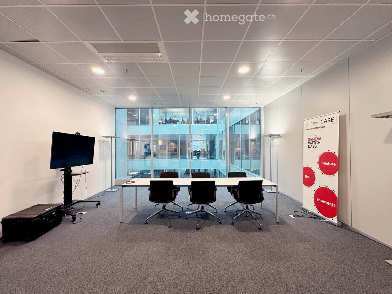 Modern office meeting room with a long table, six chairs, a large screen on a stand, and a floor lamp. Large windows provide natural light, and a banner is displayed on the wall.
