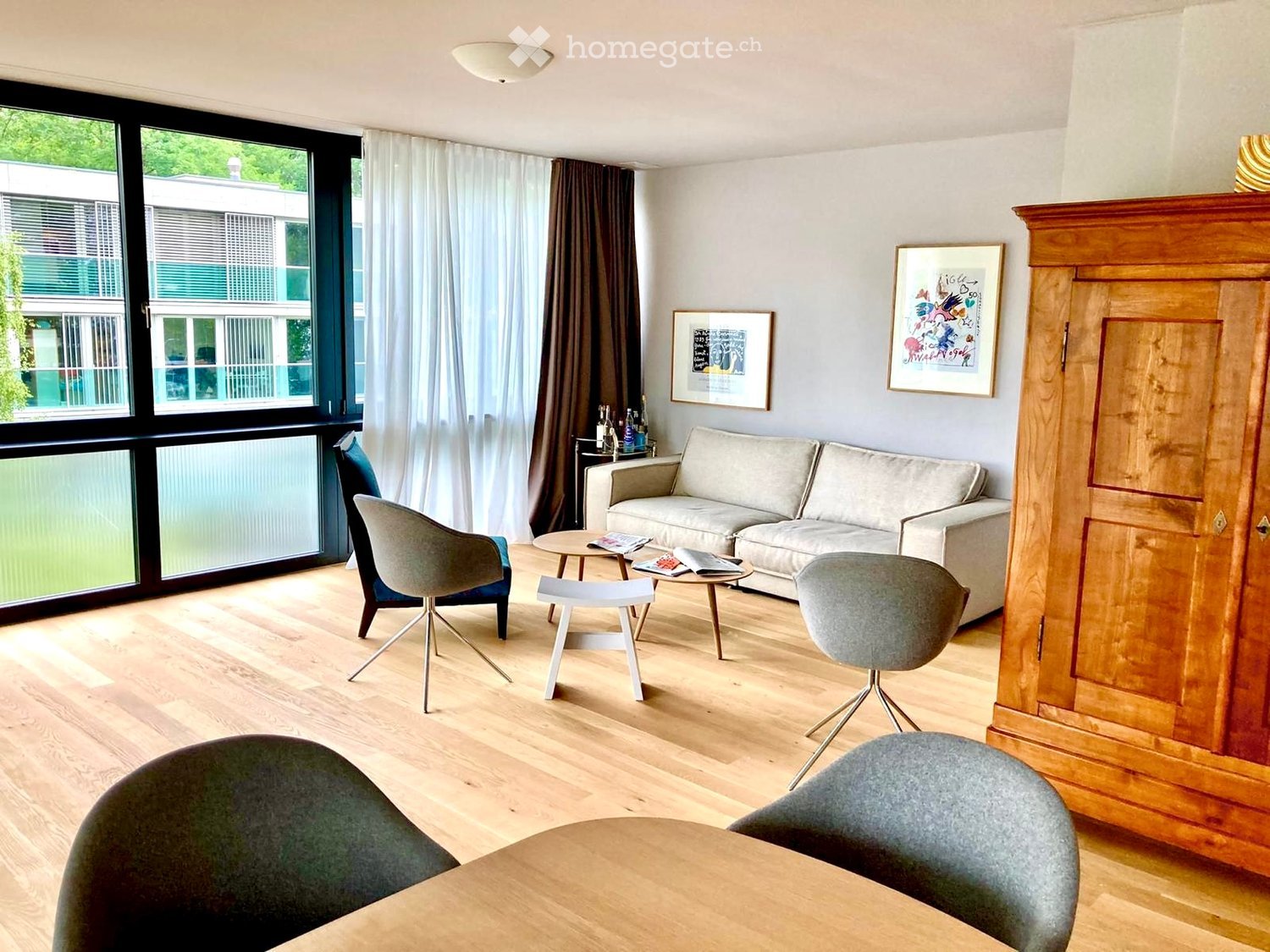 Modern living room with light wooden flooring, large windows with curtains, a sofa, chairs, a small table, and a wooden cabinet.