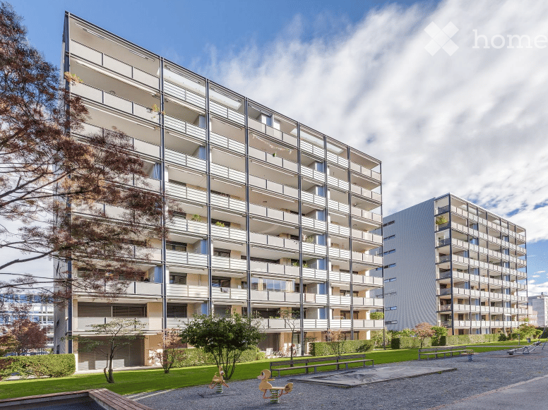 Modern multi-story apartment building with balconies and large windows, surrounded by greenery and a playground area.