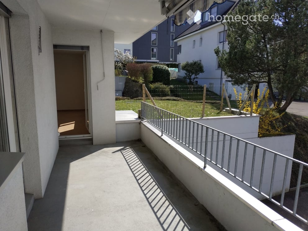 Balcony with metal railing, concrete floor, and access to an interior space. The view includes a garden area with trees and neighboring buildings.