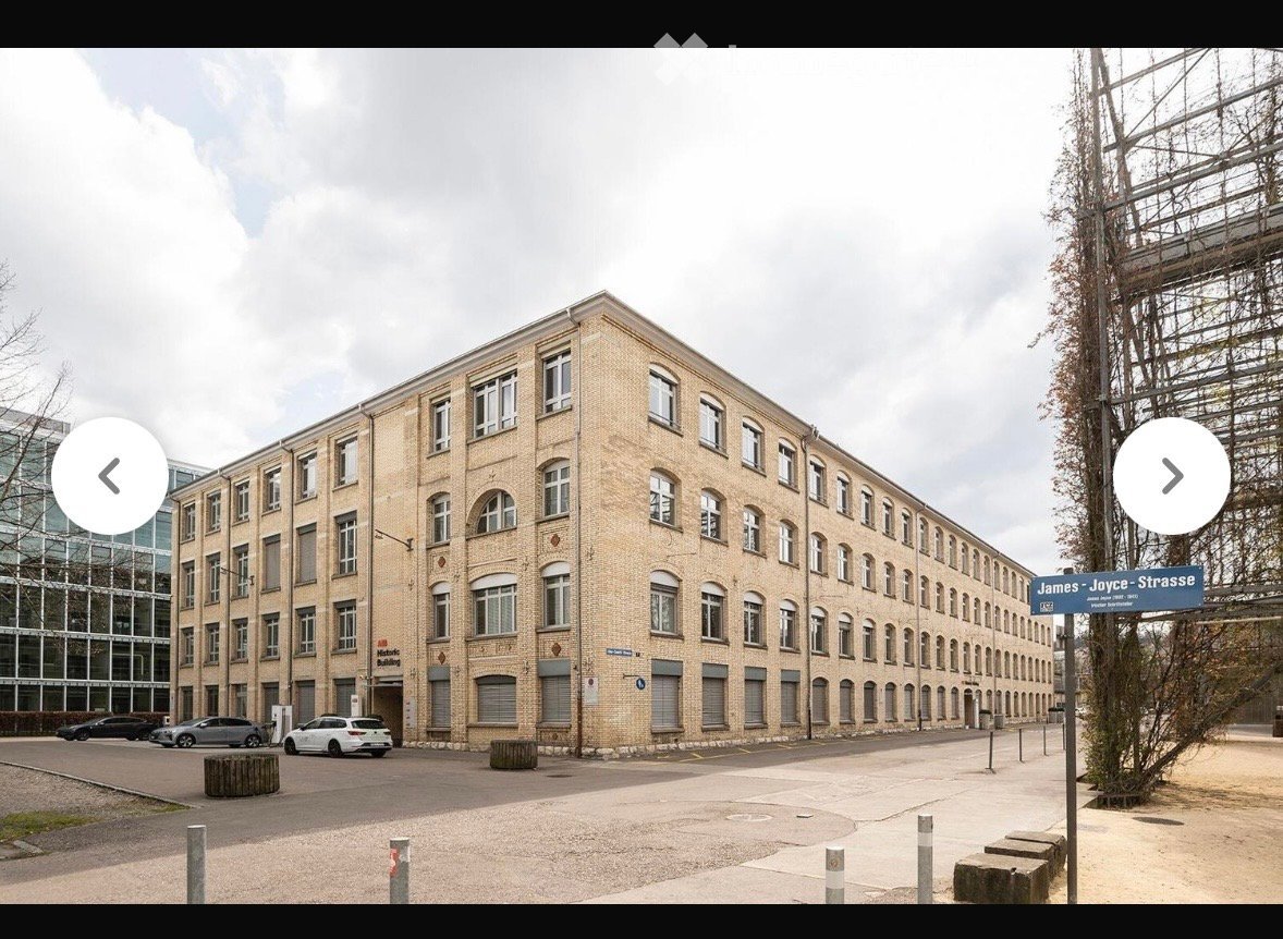 Large brick building with multiple windows and a long facade, located on a paved area with several parked cars.