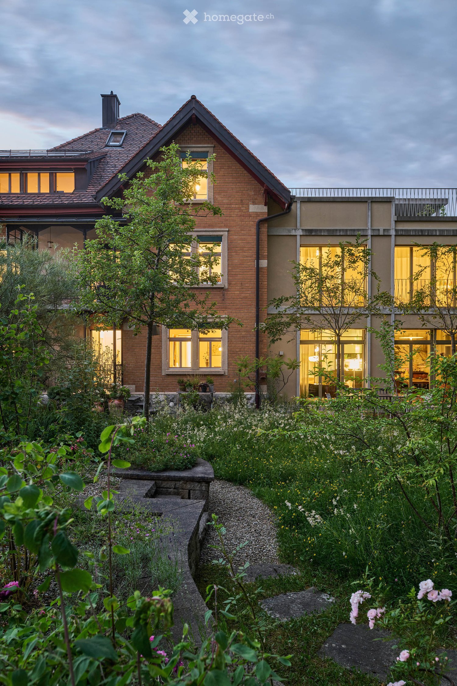 A modern two-story house with a brick facade and large windows, surrounded by a lush garden with various plants, flowers, and a stone pathway.