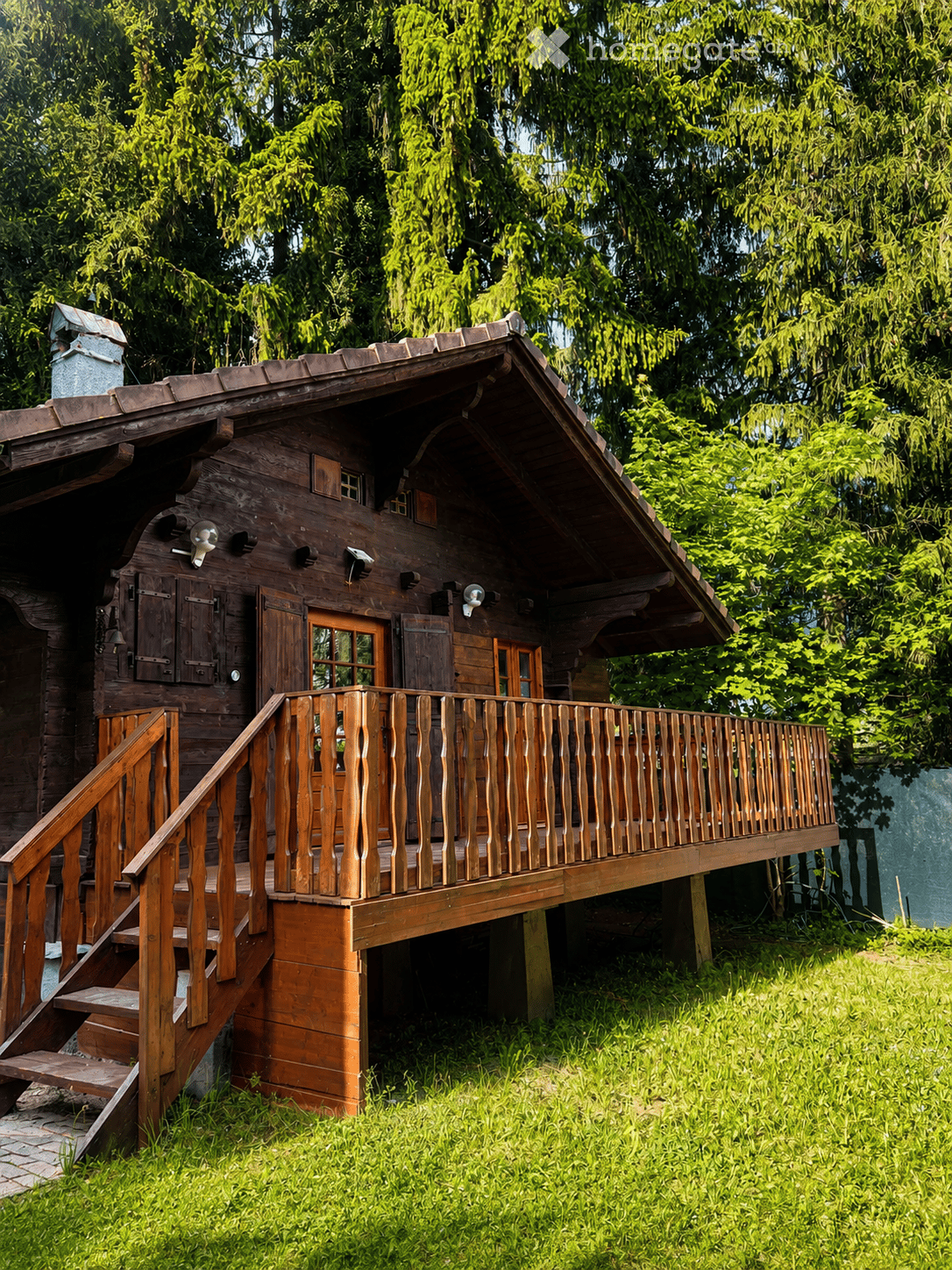 A wooden house with a raised ground floor, featuring a wooden staircase leading to a wooden balcony with a railing. The house has a sloped roof and is surrounded by lush green trees and grass.