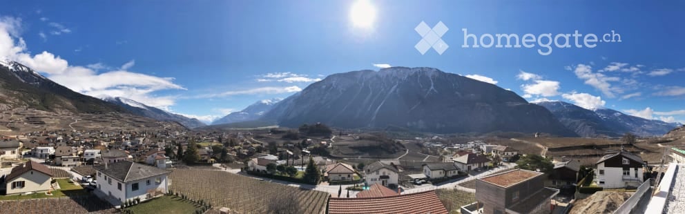 A panoramic view of a small village nestled in a valley surrounded by mountains. The village features numerous houses with varied architectural styles, surrounded by greenery and open spaces.