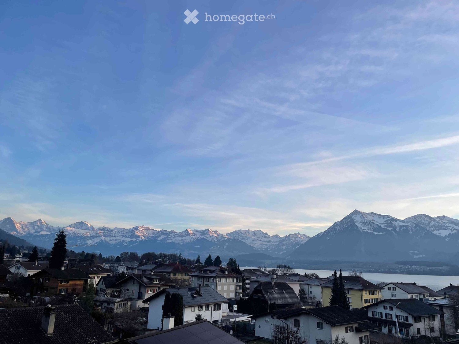 Building. A panoramic view of a town with houses and a lake, surrounded by snow-capped mountains under a cloudy sky.