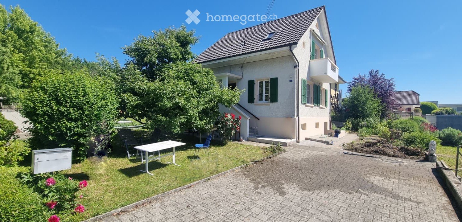 Garden area with a white table and blue chair, surrounded by greenery and a pathway leading to the house.