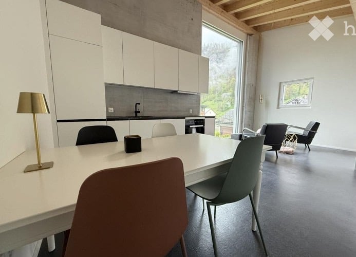 Modern, open-plan room with a dining area and kitchen. Features a white dining table with chairs, a floor lamp, and a sleek kitchen area with cabinets, a sink, and a dishwasher. Large windows offer a view of greenery outside.