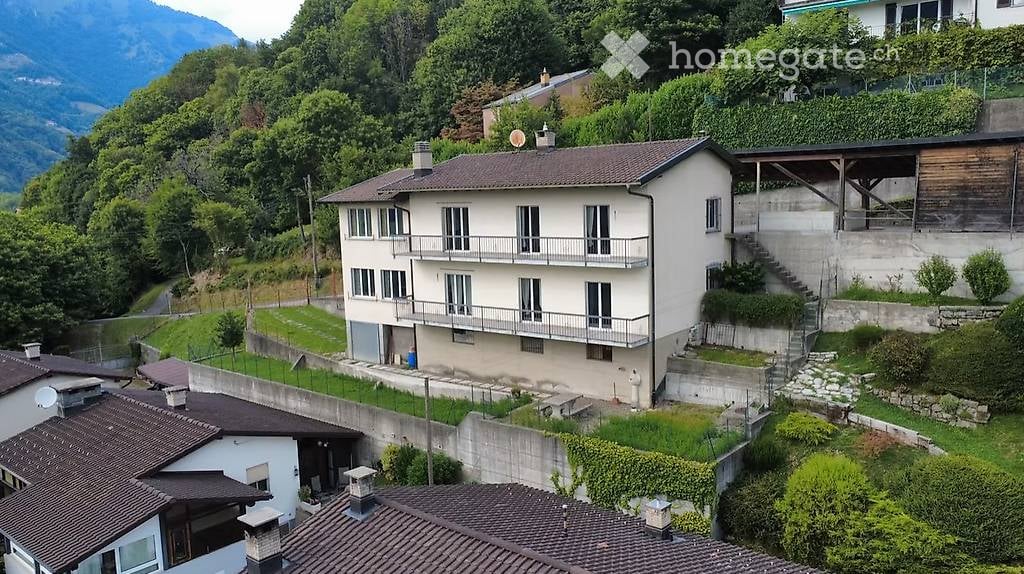 A two-story residential building with a beige facade, multiple windows, and balconies, surrounded by greenery and situated in a hilly area with visible mountains in the background.