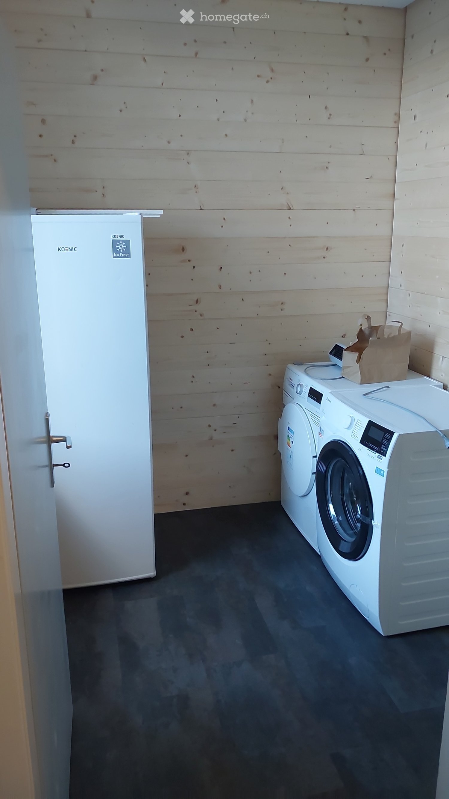 A small utility room with a refrigerator, washing machine, and a paper bag on top of the washing machine. The walls are wooden, and the floor is dark grey.