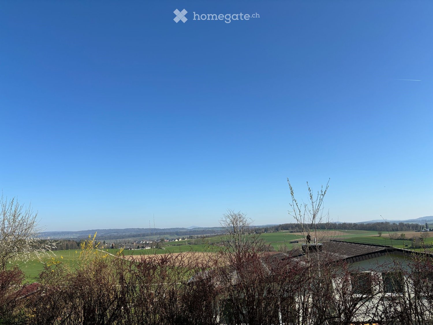A panoramic view of a rural landscape with fields, scattered houses, and distant hills under a clear blue sky.