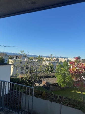 Balcony with metal railing, overlooking a cityscape with buildings, trees, and distant mountains under a clear blue sky.