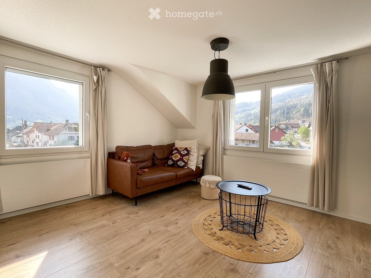 Living room with wooden flooring, large windows offering a scenic view, brown leather sofa, small round table, and decorative rug.