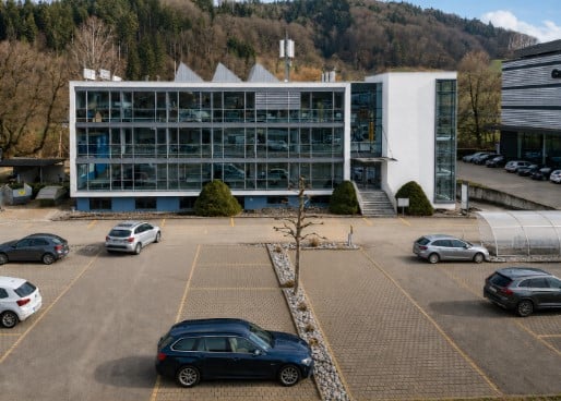 Modern office building with large glass windows and a parking lot with several cars.