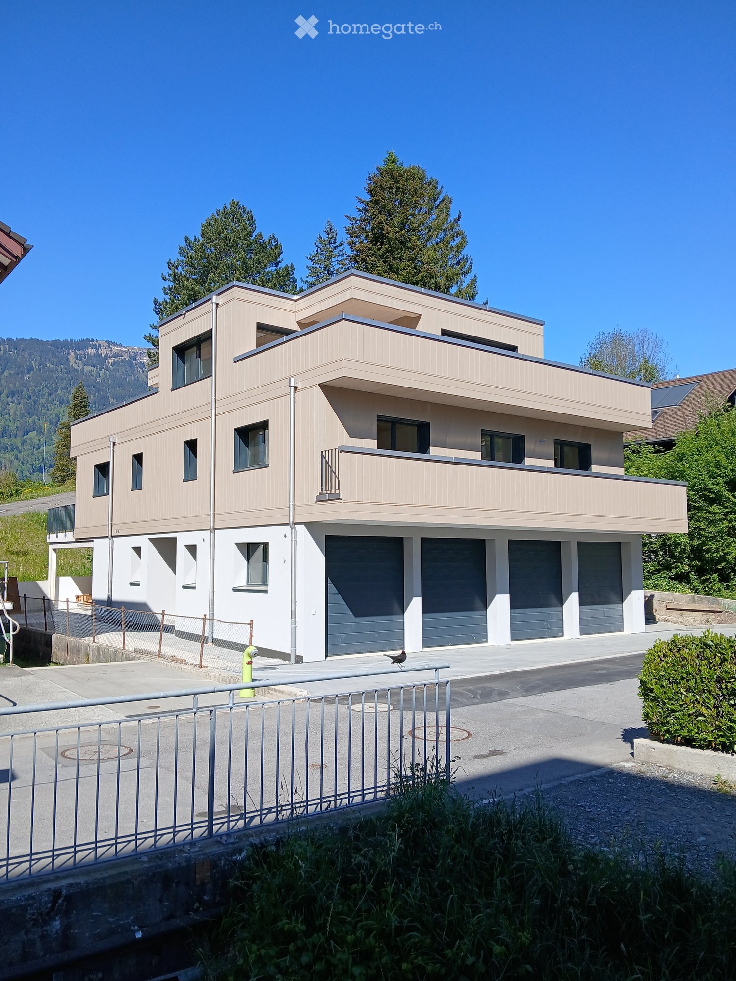 Modern two-story house with beige siding, multiple windows, and a large garage. The house features a balcony on the upper level and is surrounded by greenery and a clear blue sky.
