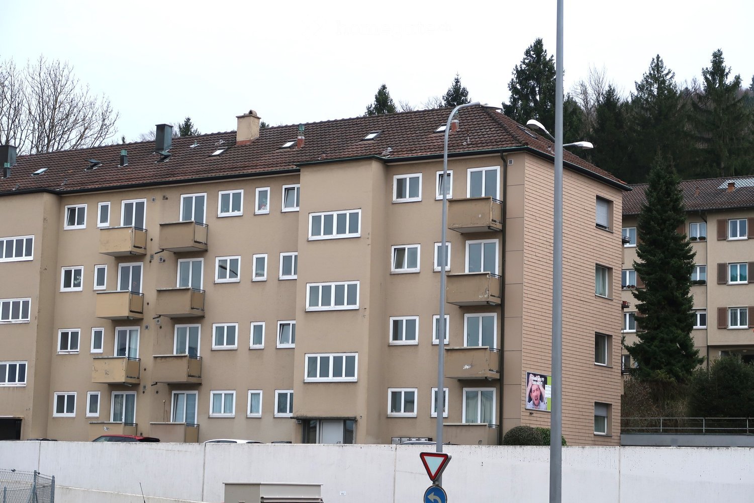 Multi-story residential building with beige facade, multiple windows, and balconies. The building has a tiled roof and is surrounded by trees and a street sign.