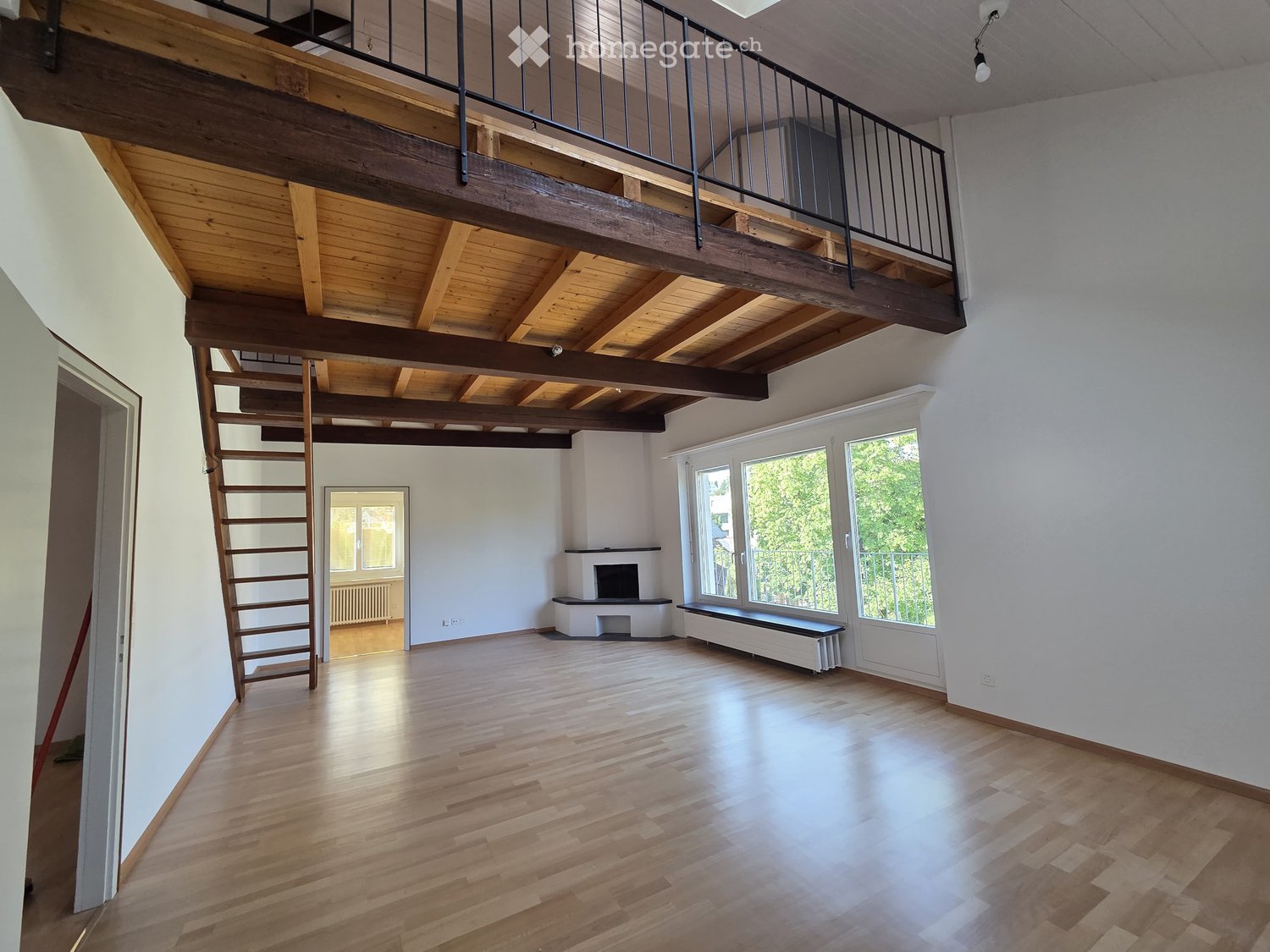 Open-plan unfurnished room with wooden flooring, exposed wooden beams, a staircase leading to a loft area, large windows, and a fireplace.