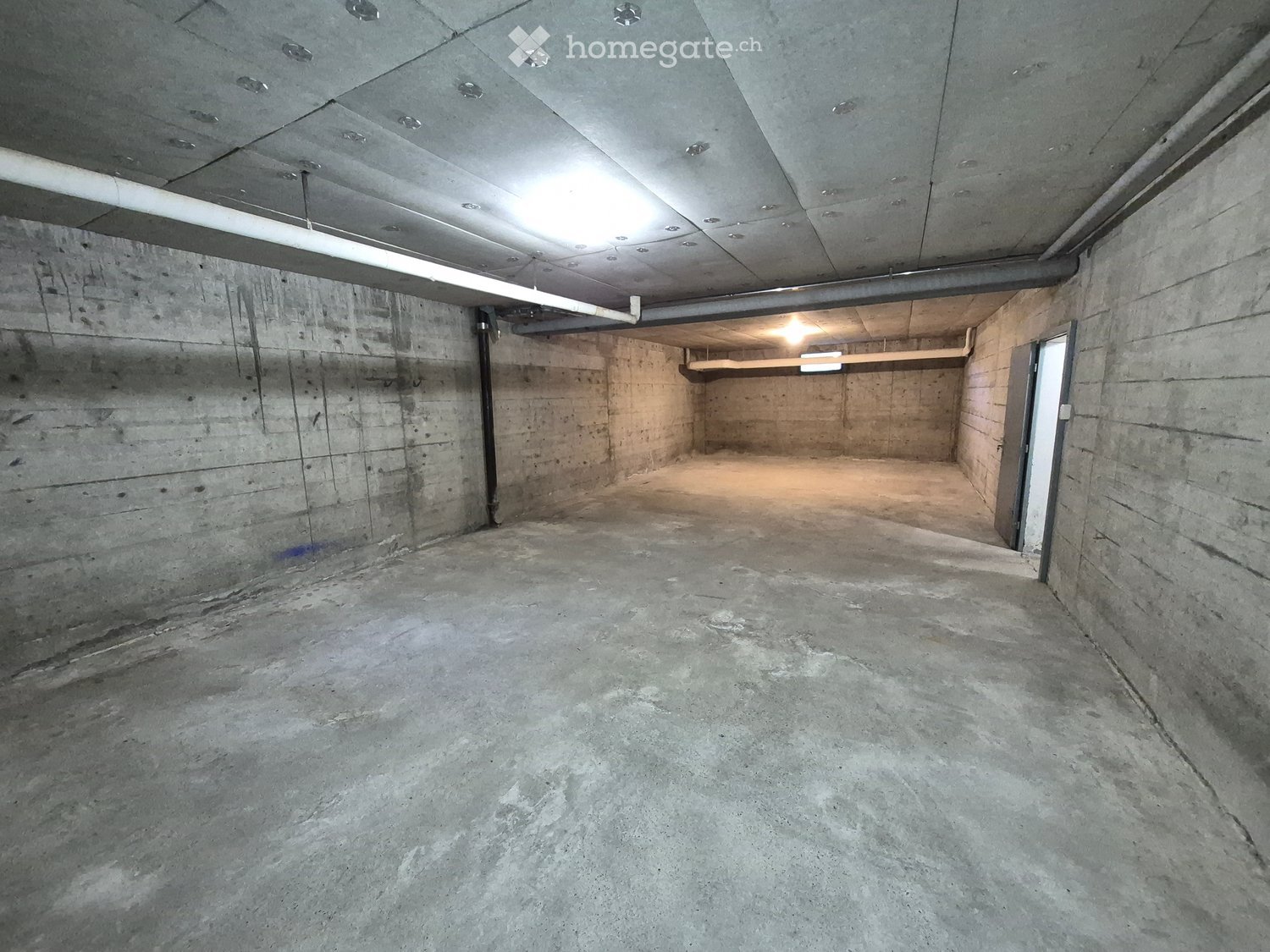 Empty underground parking space with concrete walls, floor, and ceiling, featuring a single doorway and exposed pipes.