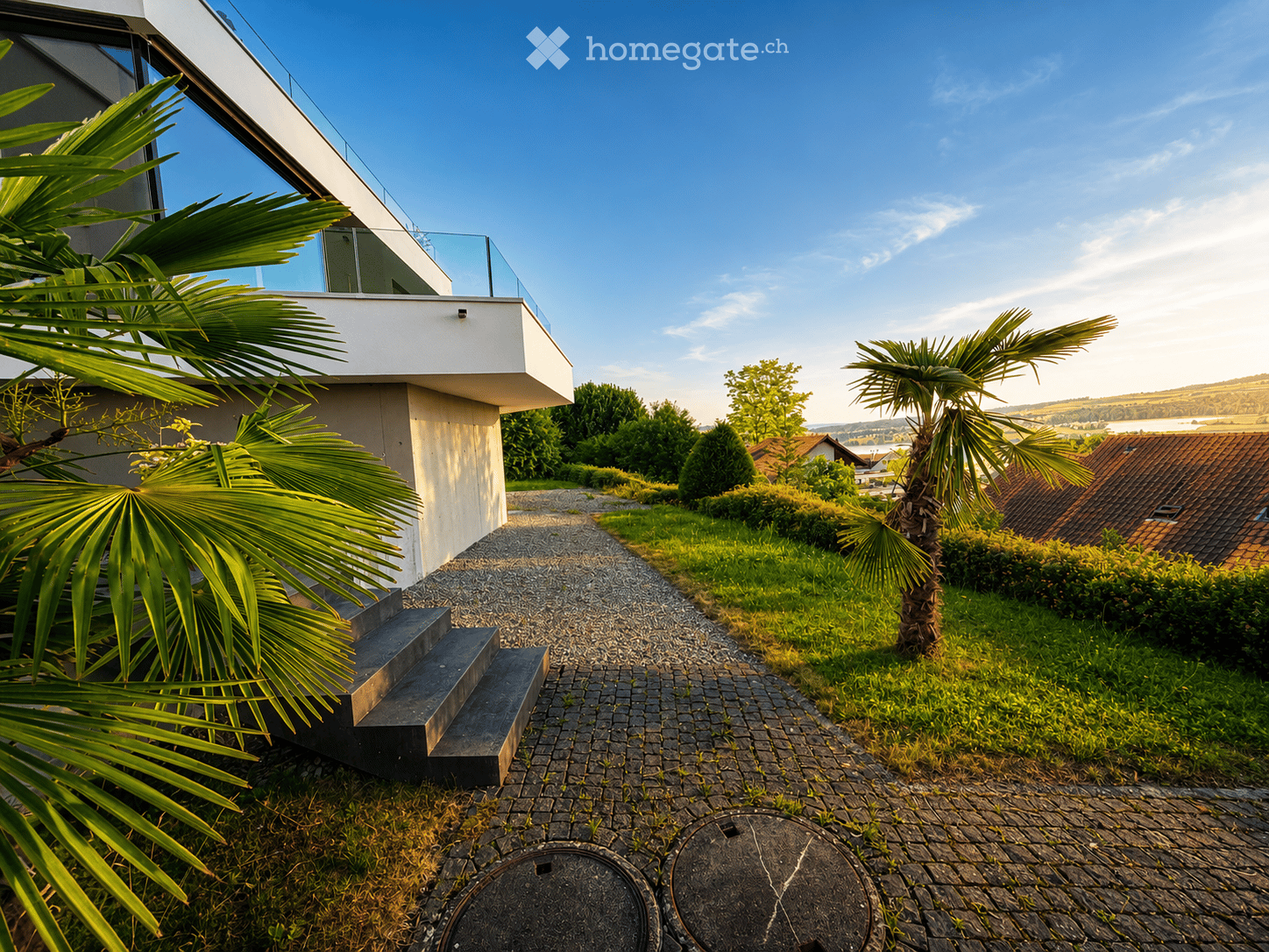 Modern house exterior with a raised ground floor, surrounded by a well-maintained garden with palm trees, gravel pathways, and a scenic view of the countryside.