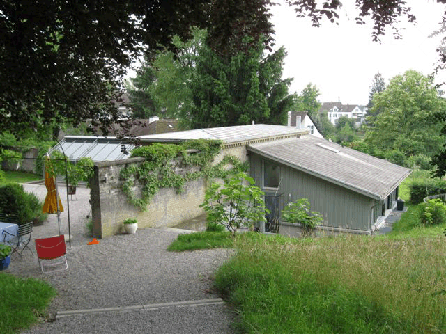 House with a raised ground floor, gray exterior, surrounded by greenery, gravel pathway, and a small garden area with a potted plant.