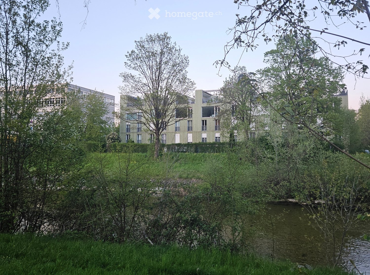 Modern multi-story building with large windows and balconies, surrounded by trees and greenery near a water body.