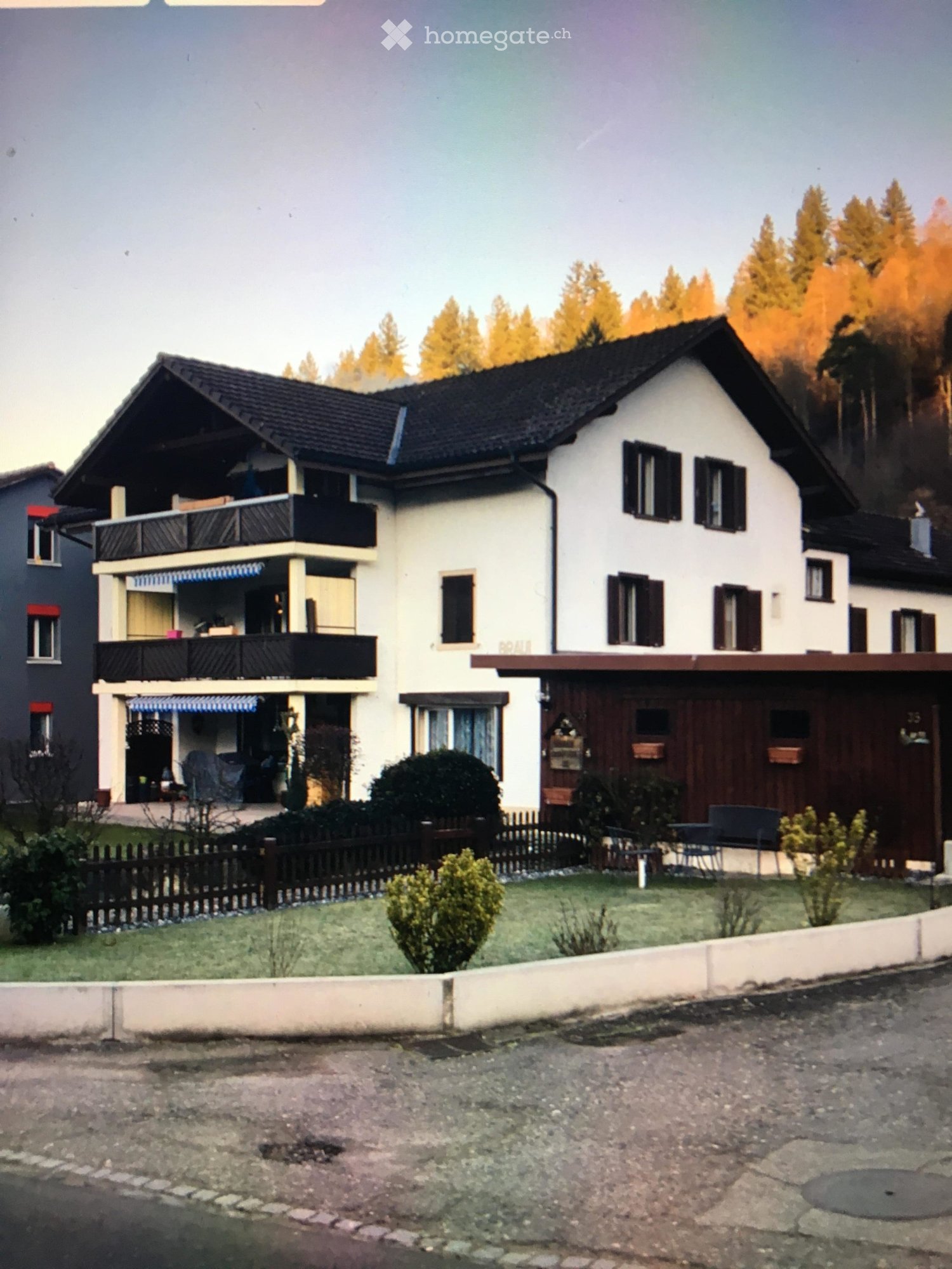 Two-story house with white walls, dark roof, multiple windows, and a balcony on the upper level. The house has a wooden garage and is surrounded by a small garden with a fence.