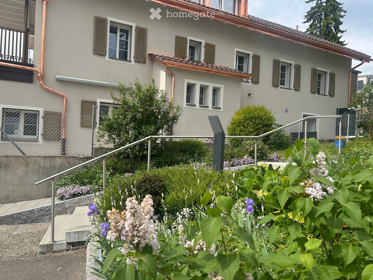 Two-story house with light gray exterior, multiple windows with shutters, and a well-maintained garden with flowers and shrubs.