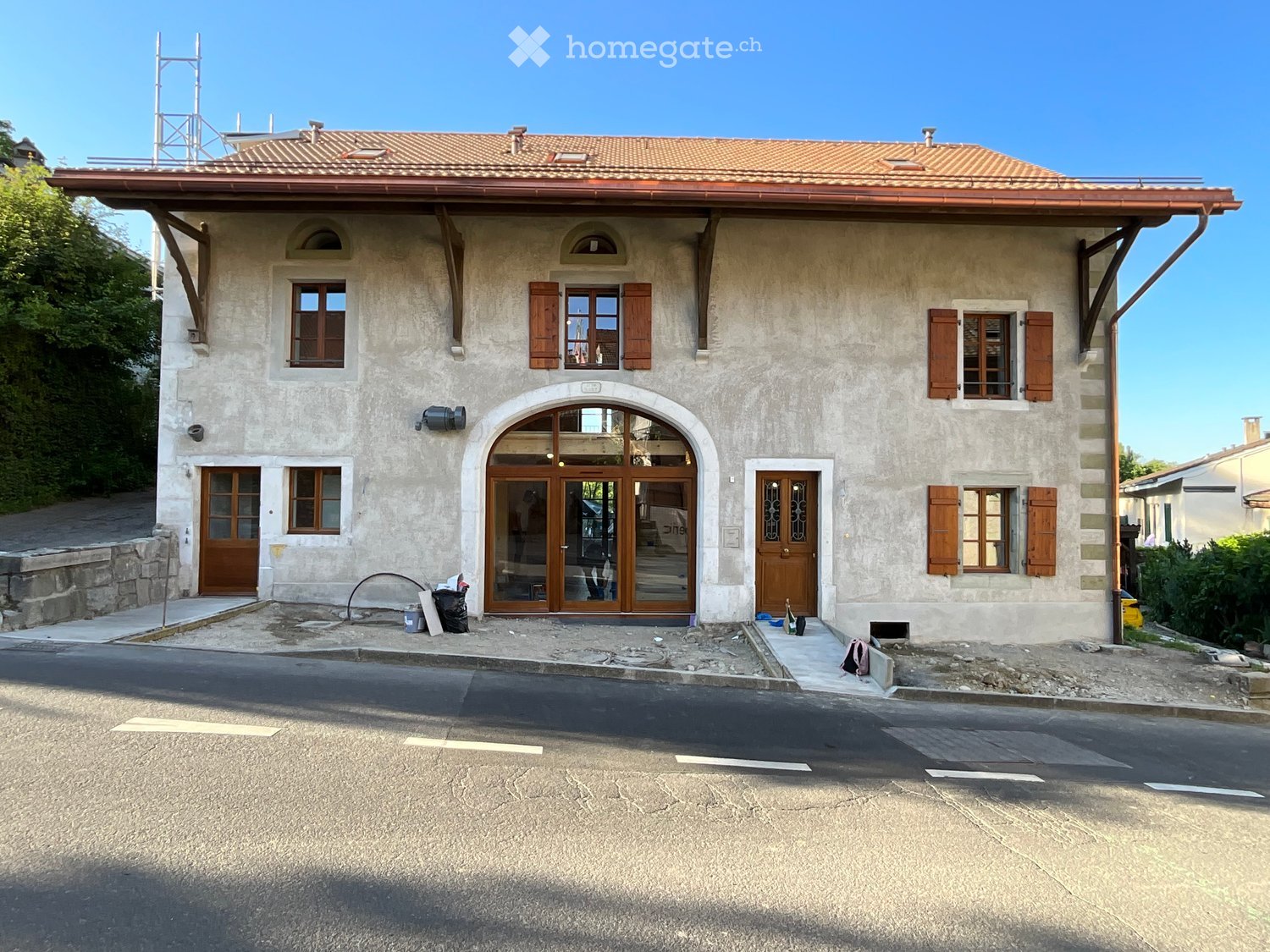 Two-story house with beige stone facade, wooden shutters, arched entrance, and a raised ground floor.