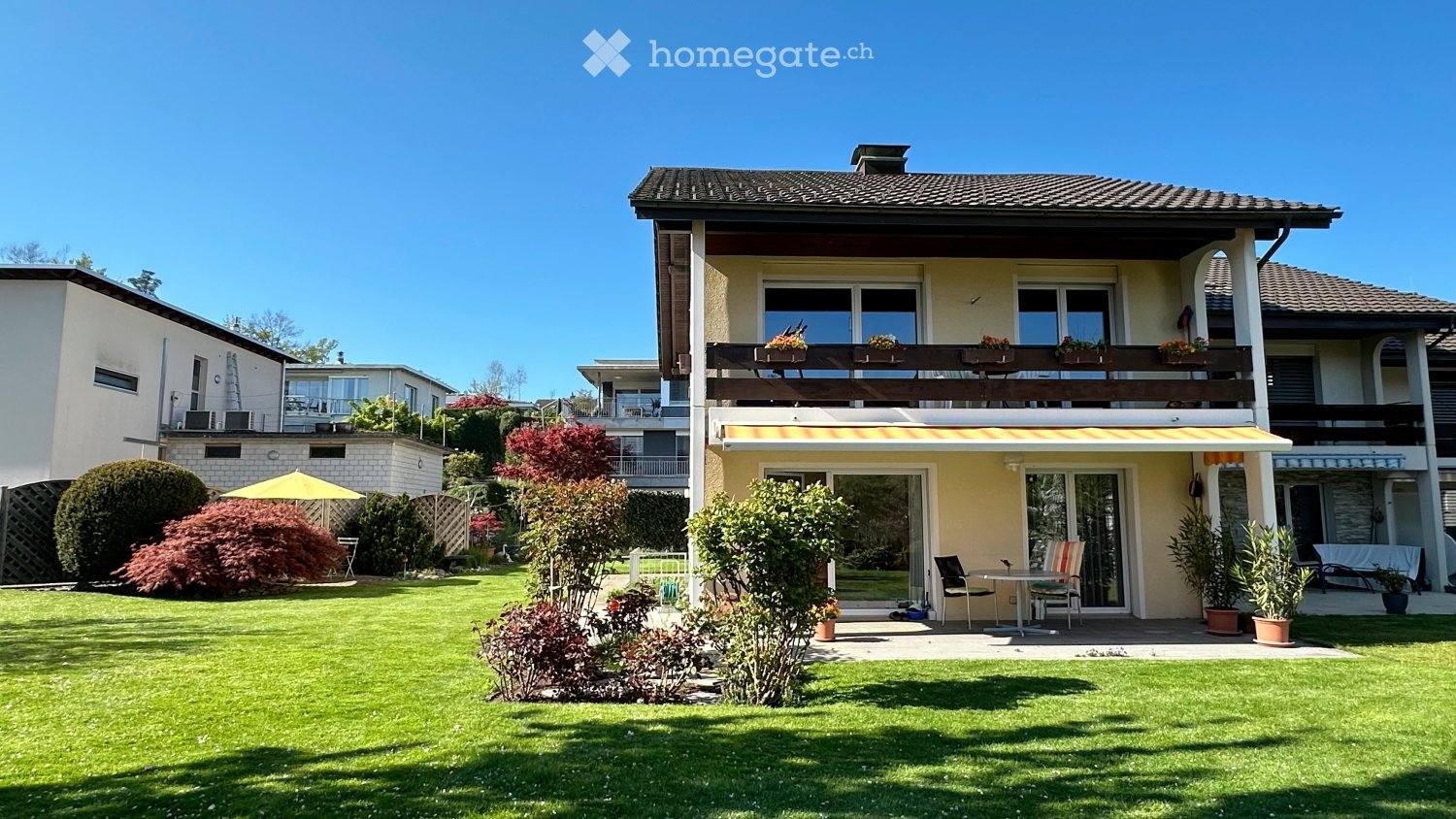 Two-story house with a balcony, surrounded by a well-maintained garden with lawn, shrubs, and potted plants. The house features large windows and a covered patio area with outdoor furniture.