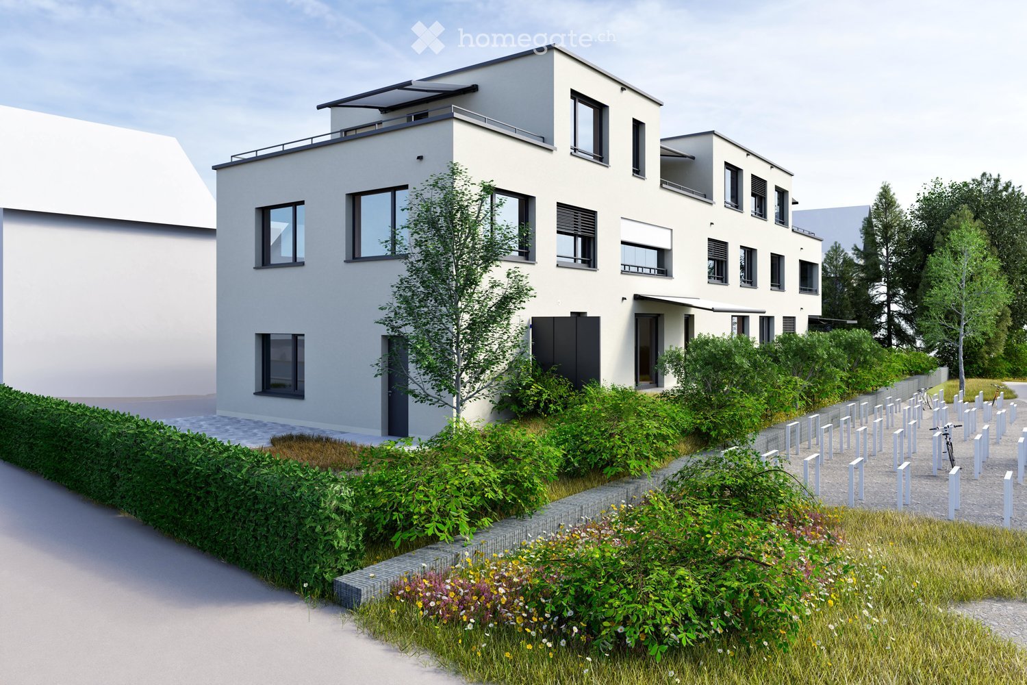 Modern two-story house with white exterior, large windows, and a balcony on the upper floor. The house is surrounded by a landscaped garden with shrubs, flowers, and a pathway.