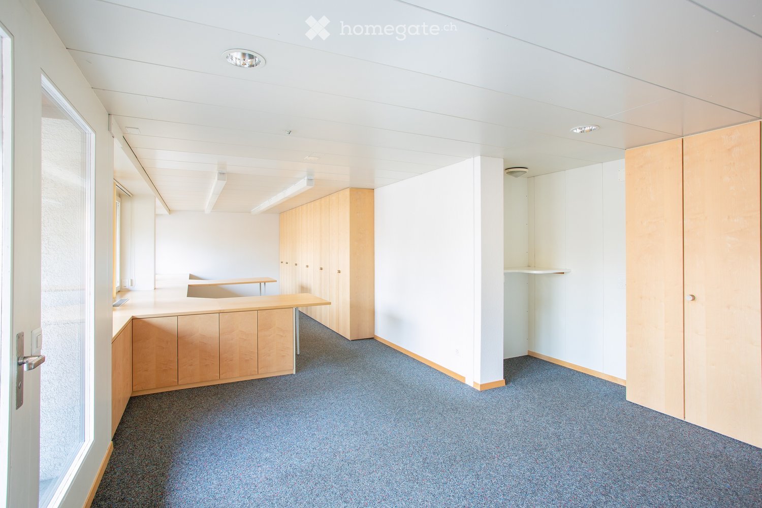 Modern, unfurnished office space with a reception desk, wooden cabinets, and shelves. The room features white walls, grey carpeting, and recessed lighting.